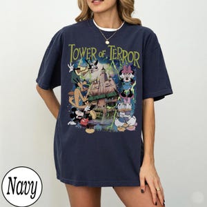 K&ouml;nnte beinhalten: Marineblaues T-Shirt mit einer Grafik von Disney-Figuren, darunter Micky Maus, Goofy und Minnie Maus, vor dem Hollywood Tower Hotel. Der Text "Tower of Terror" ist in Gr&uuml;n &uuml;ber dem Bild dargestellt.