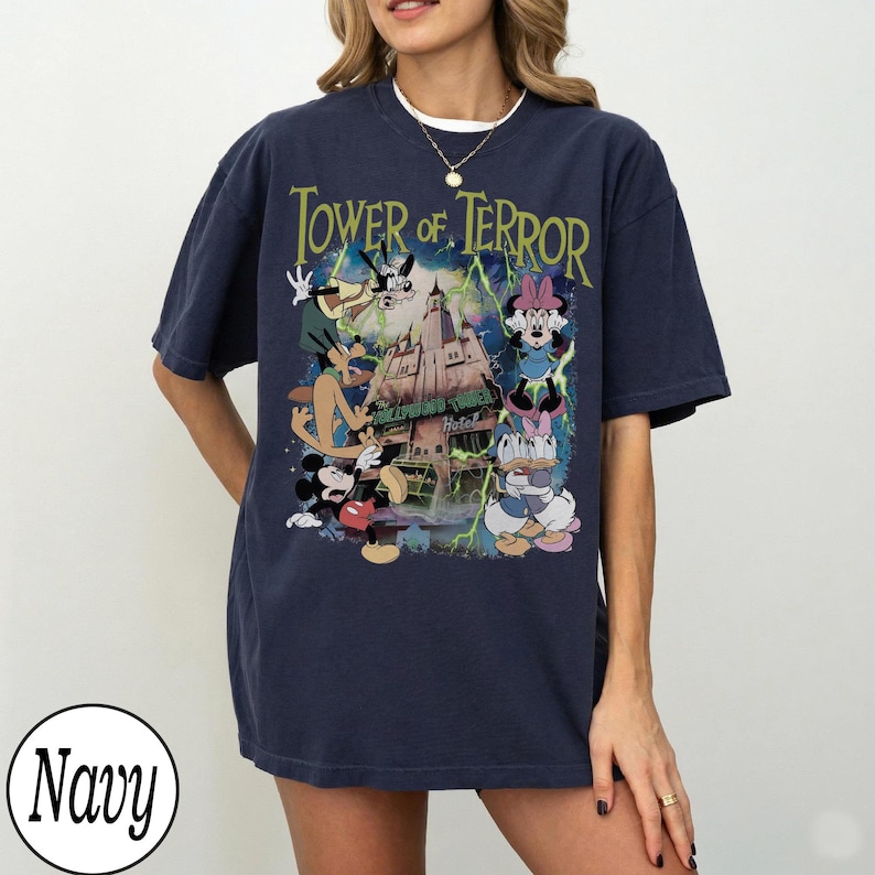 K&ouml;nnte beinhalten: Marineblaues T-Shirt mit einer Grafik von Disney-Figuren, darunter Micky Maus, Goofy und Minnie Maus, vor dem Hollywood Tower Hotel. Der Text "Tower of Terror" ist in Gr&uuml;n &uuml;ber dem Bild dargestellt.
