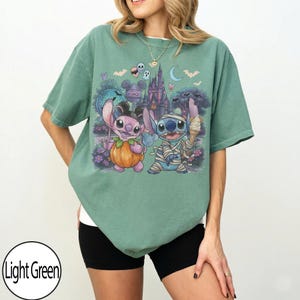 May include: A light green t-shirt featuring a graphic print of two cartoon characters dressed for Halloween. One character is dressed as a pumpkin and the other as a mummy. The background shows a castle, bats, and a crescent moon.