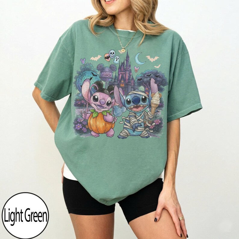 May include: A light green t-shirt featuring a graphic print of two cartoon characters dressed for Halloween. One character is dressed as a pumpkin and the other as a mummy. The background shows a castle, bats, and a crescent moon.