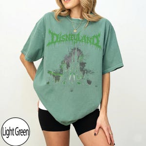 May include: Light green t-shirt with the word "DISNEYLAND" in a green, stylized font. Below the text is a graphic of a castle in shades of green and purple. The shirt is a relaxed fit.