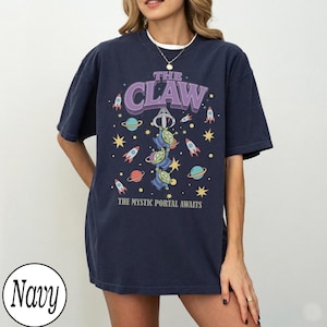 Puede incluir: Una camiseta de manga corta azul marino con un estampado gr&aacute;fico que incluye el texto THE CLAW y THE MYSTIC PORTAL AWAITS. El dise&ntilde;o muestra tres alien&iacute;genas verdes de tres ojos colgados de una garra, rodeados de cohetes, planetas y estrellas.