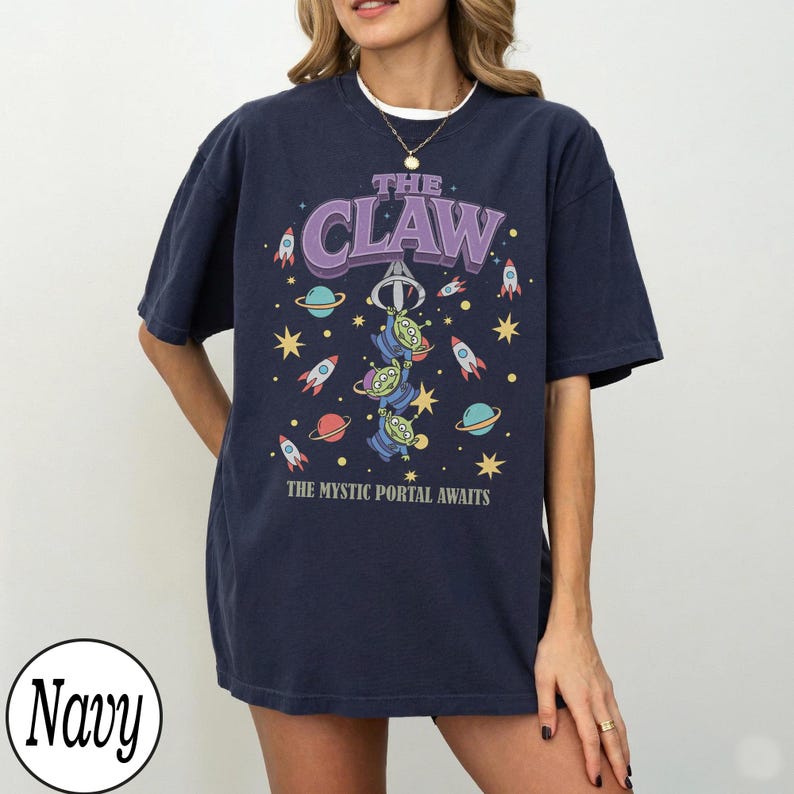 Puede incluir: Una camiseta de manga corta azul marino con un estampado gr&aacute;fico que incluye el texto THE CLAW y THE MYSTIC PORTAL AWAITS. El dise&ntilde;o muestra tres alien&iacute;genas verdes de tres ojos colgados de una garra, rodeados de cohetes, planetas y estrellas.