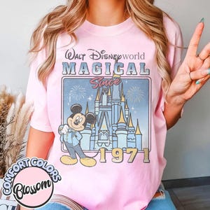 May include: Pink t-shirt with a graphic featuring Mickey Mouse, Cinderella's castle, and the text "Walt Disney World MAGICAL Since 1971." The shirt has a Comfort Colors Blossom logo.