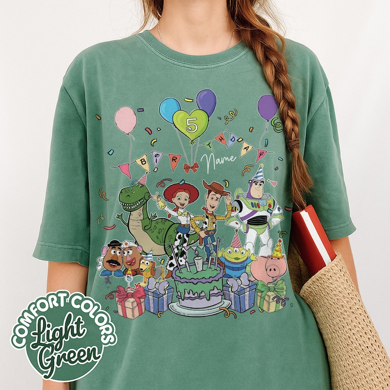 May include: Light green t-shirt featuring a colorful graphic of Toy Story characters celebrating a birthday. The design includes balloons, a cake, and the number 5. The shirt also has the words "Comfort Colors" and "Light Green" printed on it.