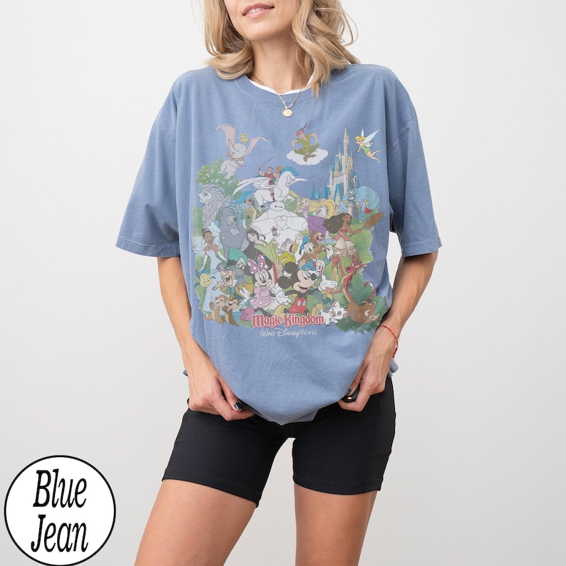May include: A light blue t-shirt featuring a colorful graphic of various Disney characters, including Mickey Mouse, Dumbo, and Tinkerbell. The text "Magic Kingdom, Walt Disney World" is printed below the characters. The shirt is paired with black shorts.