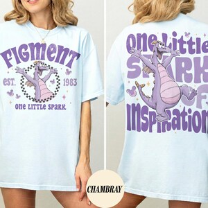 May include: Light blue t-shirt with a purple dragon graphic and text. The front of the shirt says "Figment" and "one LITTLE SPARK" with the year "1983". The back of the shirt says "one Little SPARK of Inspiration".