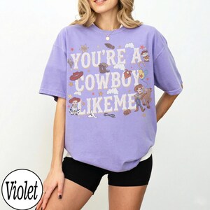 May include: A violet short-sleeve t-shirt featuring the text "YOU'RE A COWBOY LIKE ME" in white letters. The graphic includes illustrations of cartoon characters, cowboy hats, boots, stars, clouds, and a lasso, all in a muted color palette.