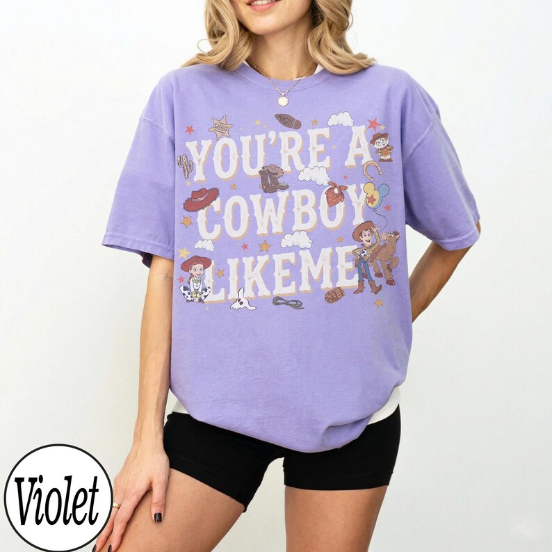 May include: A violet short-sleeve t-shirt featuring the text "YOU'RE A COWBOY LIKE ME" in white letters. The graphic includes illustrations of cartoon characters, cowboy hats, boots, stars, clouds, and a lasso, all in a muted color palette.