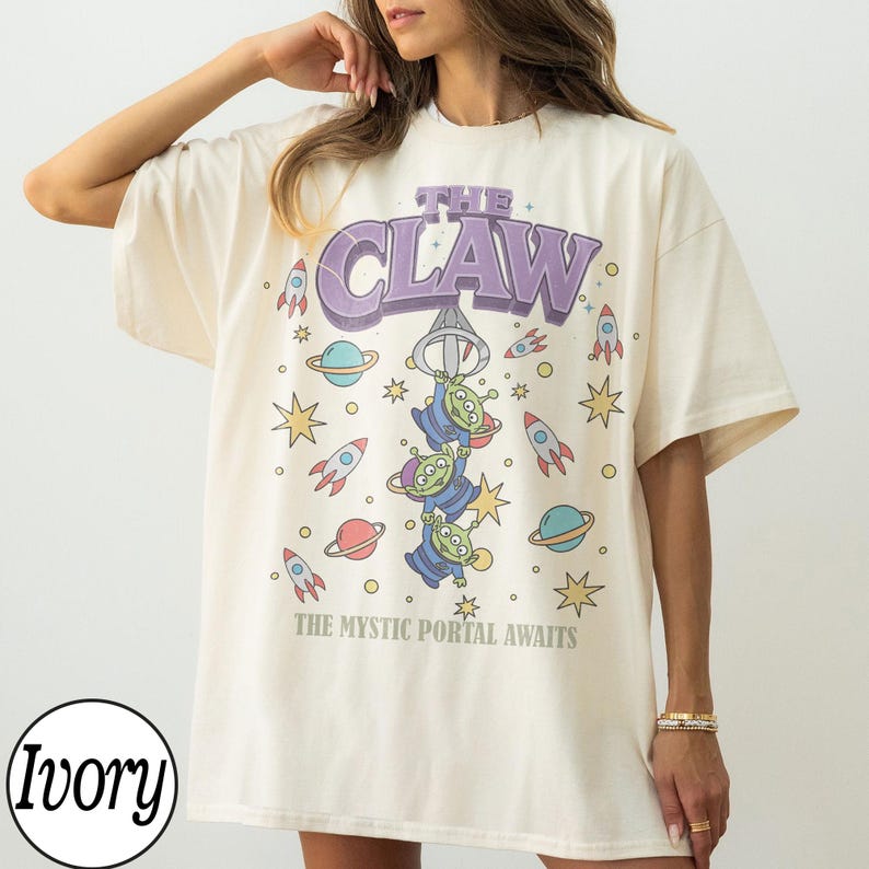 Puede incluir: Una camiseta color marfil con un estampado gr&aacute;fico de tres alien&iacute;genas verdes colgando de un gancho de m&aacute;quina de garra. El texto dice THE CLAW y THE MYSTIC PORTAL AWAITS. El dise&ntilde;o incluye cohetes, planetas y estrellas coloridos en tonos morados.