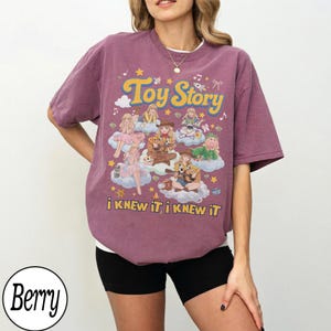 Może przedstawiać: Koszulka z kr&oacute;tkim rękawem w kolorze jagodowym z nadrukiem graficznym zawierającym tekst Toy Story oraz i KNEW IT, i KNEW IT. Wz&oacute;r przedstawia kilka ilustrowanych postaci z filmu siedzących na chmurach, otoczonych gwiazdami i nutami muzycznymi.