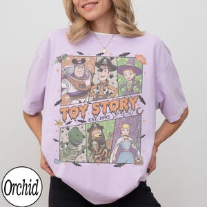 May include: A light purple t-shirt featuring a graphic print of Toy Story characters in a comic book style layout. The design includes Buzz Lightyear, Woody, Jessie, Rex, Slinky Dog, and Bo Peep with the text Toy Story Est. 1995.