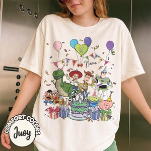 May include: Cream-colored t-shirt featuring a colorful graphic of Toy Story characters, balloons, and a birthday cake. The design includes the number 5, the word "Name", and the text "BIRTHDAY". The shirt has the "COMFORT COLORS" and "Juoy" logos.