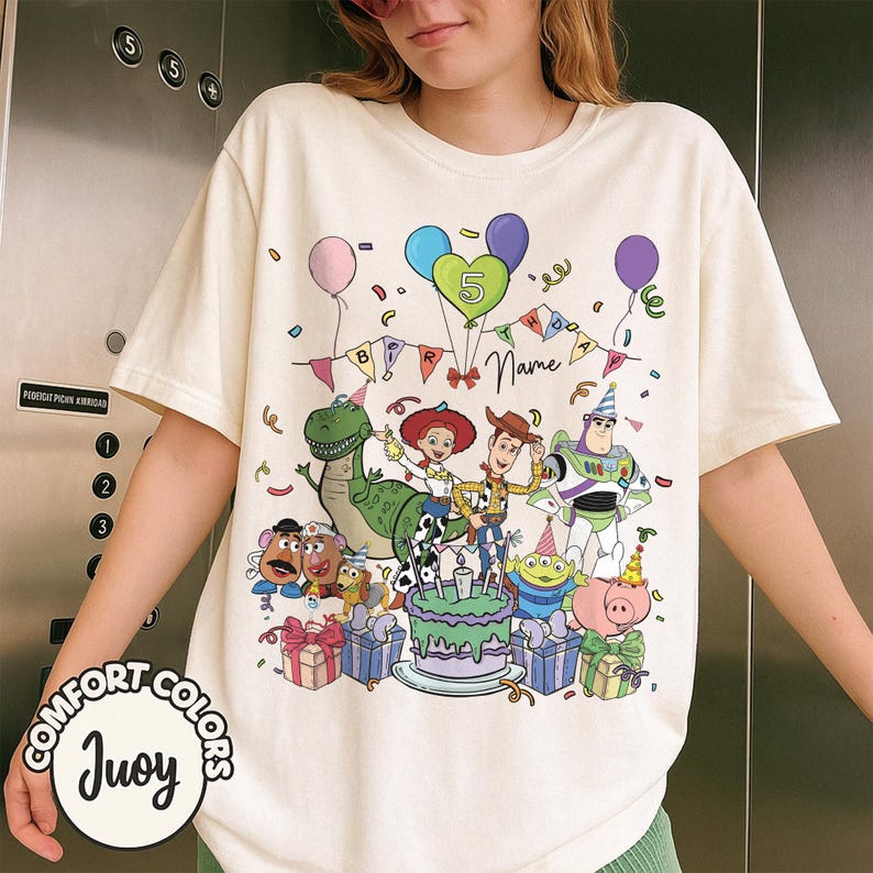 May include: Cream-colored t-shirt featuring a colorful graphic of Toy Story characters, balloons, and a birthday cake. The design includes the number 5, the word "Name", and the text "BIRTHDAY". The shirt has the "COMFORT COLORS" and "Juoy" logos.