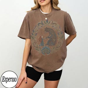 以下が含まれることがあります： 茶色のTシャツ。花輪の中で女の子を抱きしめるクマの詳細なグラフィックが特徴です。デザインには、青、緑、茶色の色合いが組み込まれています。シャツは黒いショートパンツと合わせて着用されています。「Espersso」というブランド名が下部の白い円の中にあります。