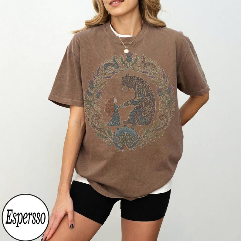 以下が含まれることがあります： 茶色のTシャツ。花輪の中で女の子を抱きしめるクマの詳細なグラフィックが特徴です。デザインには、青、緑、茶色の色合いが組み込まれています。シャツは黒いショートパンツと合わせて着用されています。「Espersso」というブランド名が下部の白い円の中にあります。