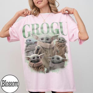 Puede incluir: Camiseta rosa claro con la palabra "GROGU" en verde y varias ilustraciones del personaje Grogu. La camiseta tiene mangas cortas y un corte holgado. El logo de Blossom est&aacute; en la esquina inferior izquierda.