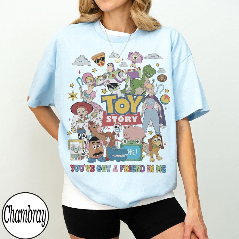 May include: A light blue t-shirt featuring a colorful graphic print of Toy Story characters including Buzz Lightyear, Woody, Jessie, and others. The text "TOY STORY" and "YOU'VE GOT A FRIEND IN ME" is printed in the center with various small icons.