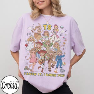 May include: A light purple t-shirt featuring a graphic print of multiple illustrated characters in various outfits, the text "TS" at the top, and the text "i KNEW IT, i KNEW YOU" at the bottom. The design includes musical notes, a rocket, and a ball.