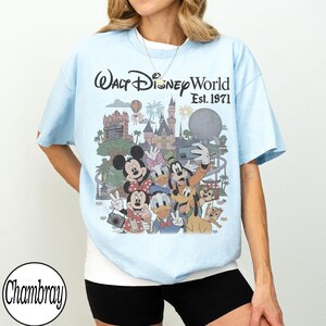 May include: A light blue t-shirt featuring a graphic print of Walt Disney World Est. 1971. The design includes illustrations of Mickey Mouse, Minnie Mouse, Donald Duck, Daisy Duck, Goofy, Pluto, and Chip 'n Dale in front of park landmarks.