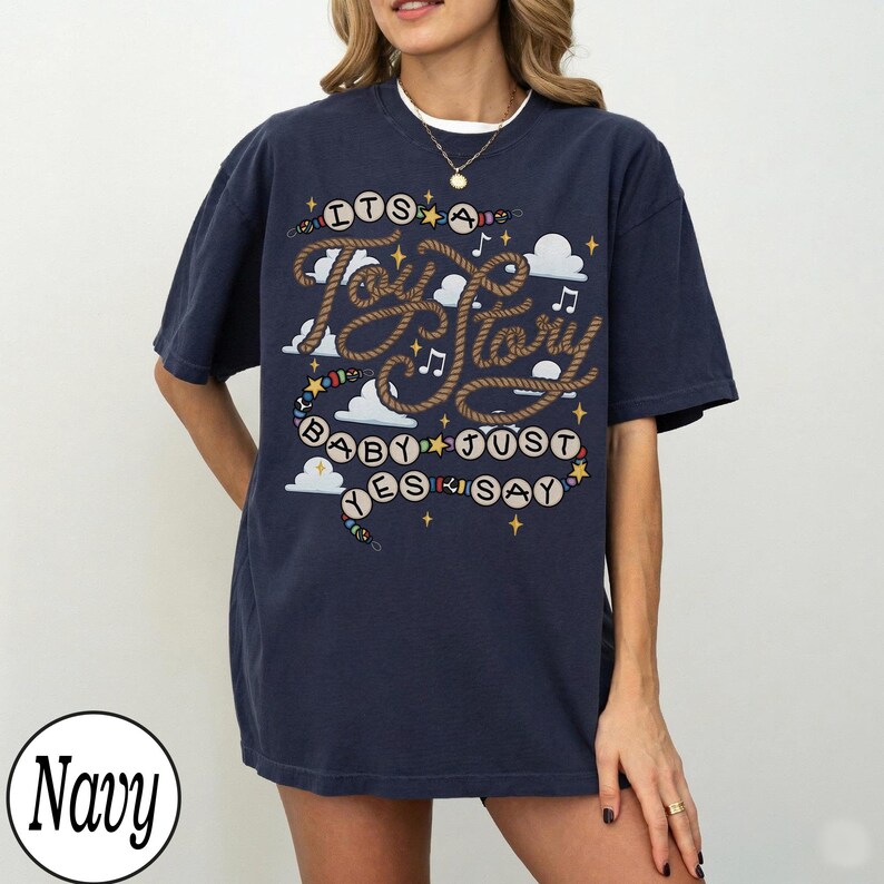 May include: A navy blue short-sleeve t-shirt featuring a graphic design with the text "ITS A Toy Story BABY JUST YES SAY". The design includes rope-style lettering, musical notes, white clouds, and small yellow stars on a dark background.