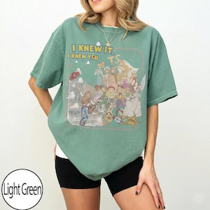 May include: A light green short-sleeve t-shirt featuring a graphic print of various animated characters and the text "I KNEW IT, I KNEW YOU" along with "TS". The design includes a person playing a guitar, a sheriff star, and a rocket ship.