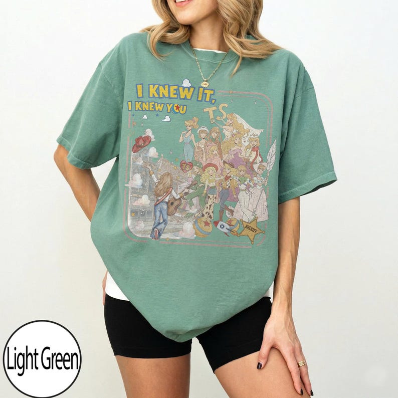 May include: A light green short-sleeve t-shirt featuring a graphic print of various animated characters and the text "I KNEW IT, I KNEW YOU" along with "TS". The design includes a person playing a guitar, a sheriff star, and a rocket ship.