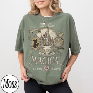 May include: Sage green t-shirt with a vintage-style graphic featuring a castle, teacup, and Mickey Mouse silhouette. The text reads "The Most Magical Place on Earth". The shirt has a relaxed fit.