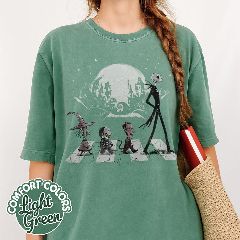 Peut inclure: T-shirt vert clair avec un motif de personnages de L'&Eacute;trange No&euml;l de Monsieur Jack traversant un passage pi&eacute;ton, avec une pleine lune et une ville effrayante en arri&egrave;re-plan. Le texte "COMFORT COLORS Light Green" est imprim&eacute; en bas &agrave; gauche.