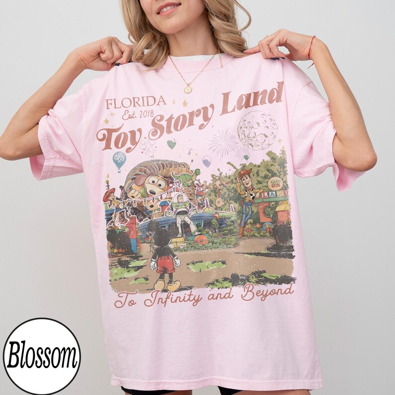 May include: A pale pink t-shirt with a graphic featuring characters from Toy Story and the text "Toy Story Land" and "To Infinity and Beyond". The shirt also has the text "Florida Est. 2018".