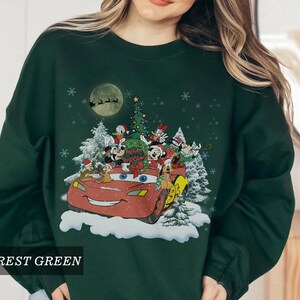 K&ouml;nnte beinhalten: Forstgr&uuml;nes Sweatshirt mit festlichem Aufdruck. Das Design zeigt ein rotes Auto mit Disney-Figuren, einen Weihnachtsbaum und den Text "Merry Christmas". Schneeflocken und ein Vollmond vervollst&auml;ndigen die Urlaubsszene.