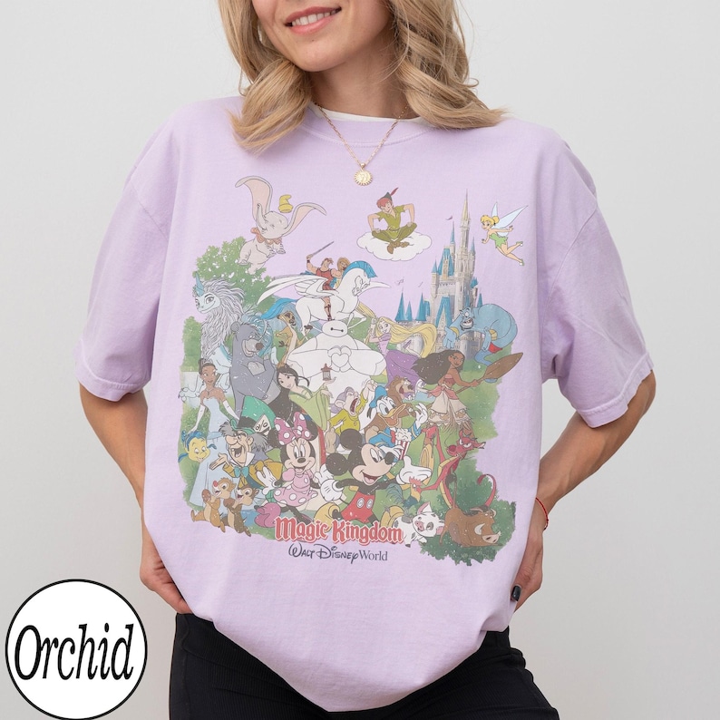 May include: A light purple t-shirt featuring a colorful graphic of various Disney characters, including Mickey Mouse, Dumbo, and Peter Pan. The text "Magic Kingdom Walt Disney World" is printed below the characters. The shirt has short sleeves and a crew neck.