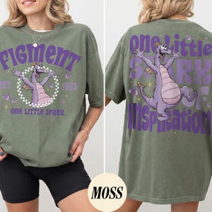 May include: Olive green t-shirt with a purple graphic of a dragon and the text "Figment" and "One Little Spark". The back of the shirt features the same dragon graphic and the text "One Little Spark of Inspiration".