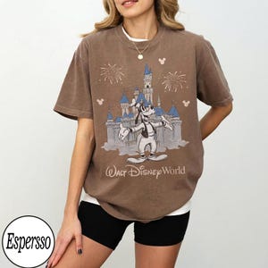 May include: Brown t-shirt with a graphic of Goofy in front of Cinderella's castle, fireworks, and the text "Walt Disney World." The shirt has a vintage style and a loose fit.