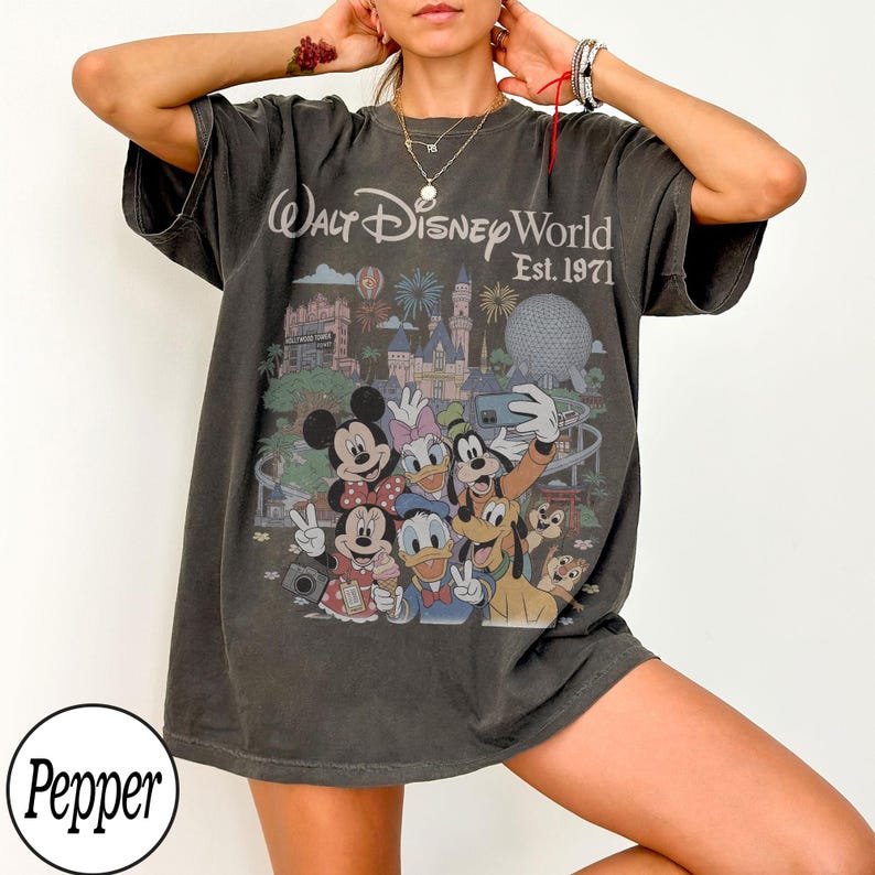 May include: A charcoal gray short-sleeve t-shirt featuring a graphic print of Disney characters including Mickey Mouse, Minnie Mouse, Donald Duck, Daisy Duck, Goofy, and Pluto. The text reads Walt Disney World Est. 1971 with a castle and park icons.