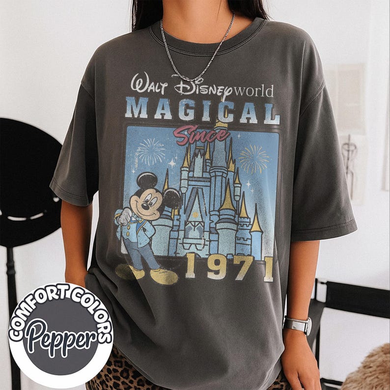 May include: Dark gray t-shirt featuring a graphic of Mickey Mouse in front of Cinderella's castle. The shirt reads "Walt Disney World Magical Since 1971". The shirt has a vintage, distressed look.