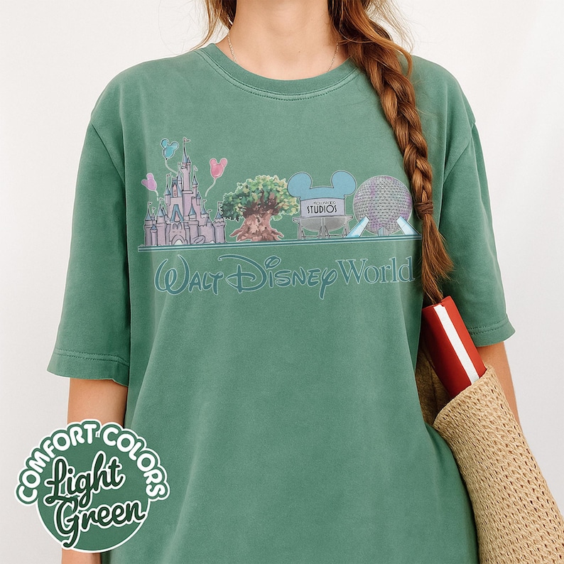 Puede incluir: Camiseta verde claro Comfort Colors con un gr&aacute;fico de Walt Disney World. El dise&ntilde;o incluye un castillo, un &aacute;rbol, orejas de Mickey Mouse y una esfera. Las palabras "Walt Disney World" est&aacute;n escritas debajo.
