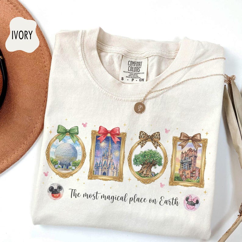 K&ouml;nnte beinhalten: Elfenbeinfarbenes T-Shirt mit vier gerahmten Illustrationen von Themenpark-Wahrzeichen, die jeweils mit einer Schleife verziert sind. Das Shirt enth&auml;lt den Text "The most magical place on Earth" und ist mit einer goldenen Halskette versehen.