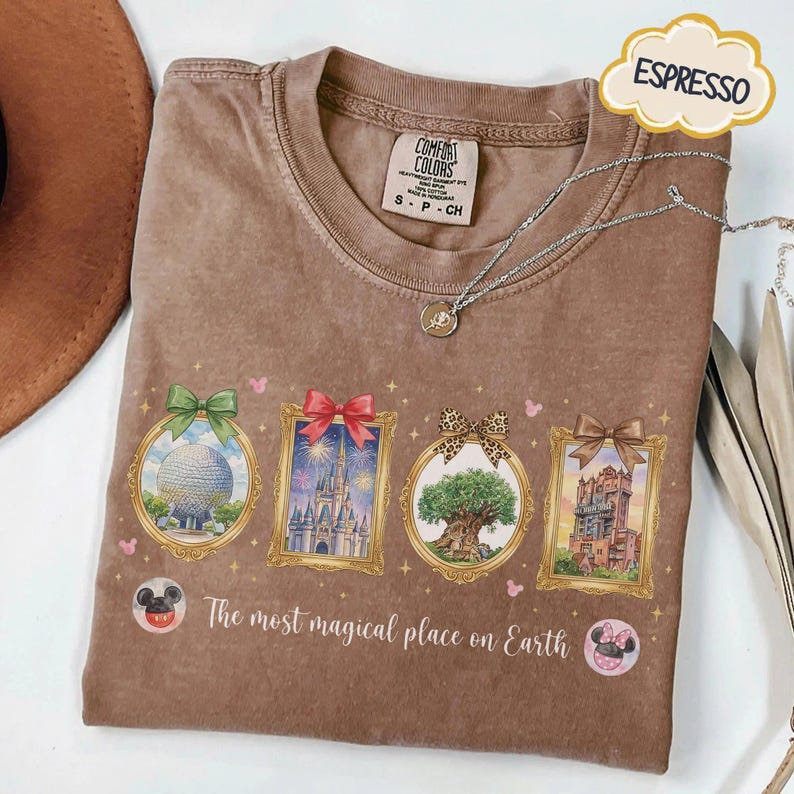 K&ouml;nnte beinhalten: Braunes T-Shirt mit vier gerahmten Illustrationen von Themenpark-Wahrzeichen. Jeder Rahmen hat ein anderes Bogendesign. Der Text "The most magical place on Earth" ist darunter gedruckt.
