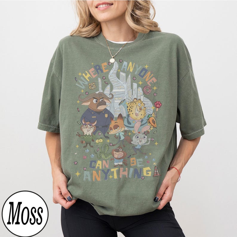 May include: Olive green t-shirt with a cartoon graphic and the text "WHERE ANYONE CAN BE ANYTHING". The design features animal characters and a cityscape. The shirt has short sleeves and a loose fit. The t-shirt is a relaxed fit.