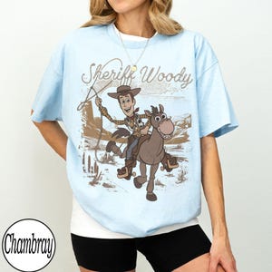 May include: Light blue t-shirt featuring a graphic of Sheriff Woody riding a horse with the text "Sheriff Woody". The design is in brown and tan tones, set against a desert backdrop. The shirt has short sleeves.