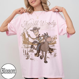 May include: A light pink t-shirt featuring a graphic of Sheriff Woody riding a horse, with the text "Sheriff Woody" above the image. The design is in shades of brown, beige, and tan. The shirt has short sleeves and a relaxed fit.