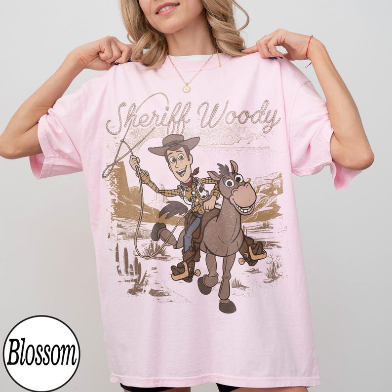 May include: A light pink t-shirt featuring a graphic of Sheriff Woody riding a horse, with the text "Sheriff Woody" above the image. The design is in shades of brown, beige, and tan. The shirt has short sleeves and a relaxed fit.