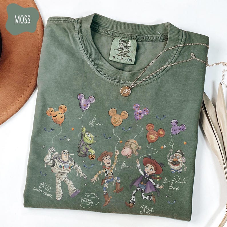 Pode incluir: Camiseta verde musgo com os personagens de desenhos animados Buzz Lightyear, Woody, Jessie, Sr. Cabe&ccedil;a de Batata e um Alien, cada um segurando bal&otilde;es em forma de Mickey Mouse. A camisa tem a etiqueta da marca Comfort Colors e um colar de ouro. A palavra "Moss" est&aacute; no canto superior esquerdo.