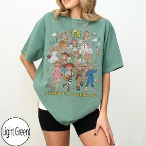 May include: A light green short-sleeve t-shirt featuring a graphic print of multiple cartoon characters in various outfits. The text "TS" appears at the top, and the text "i knew it, i knew you" is printed at the bottom of the graphic design.