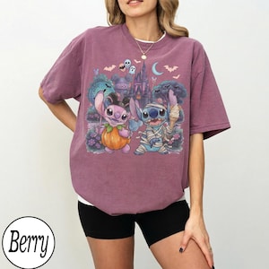 May include: A berry-colored short-sleeve t-shirt featuring a graphic print of two blue and pink alien characters dressed in Halloween costumes, one as a pumpkin and one as a mummy, with a castle, bats, and ghosts in the background.