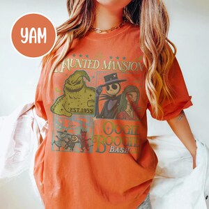 May include: Orange t-shirt featuring characters from The Nightmare Before Christmas. The design includes images of Jack Skellington, Sally, and Oogie Boogie, with text that reads "The Haunted Mansion" and "Oogie Boogie Bash."