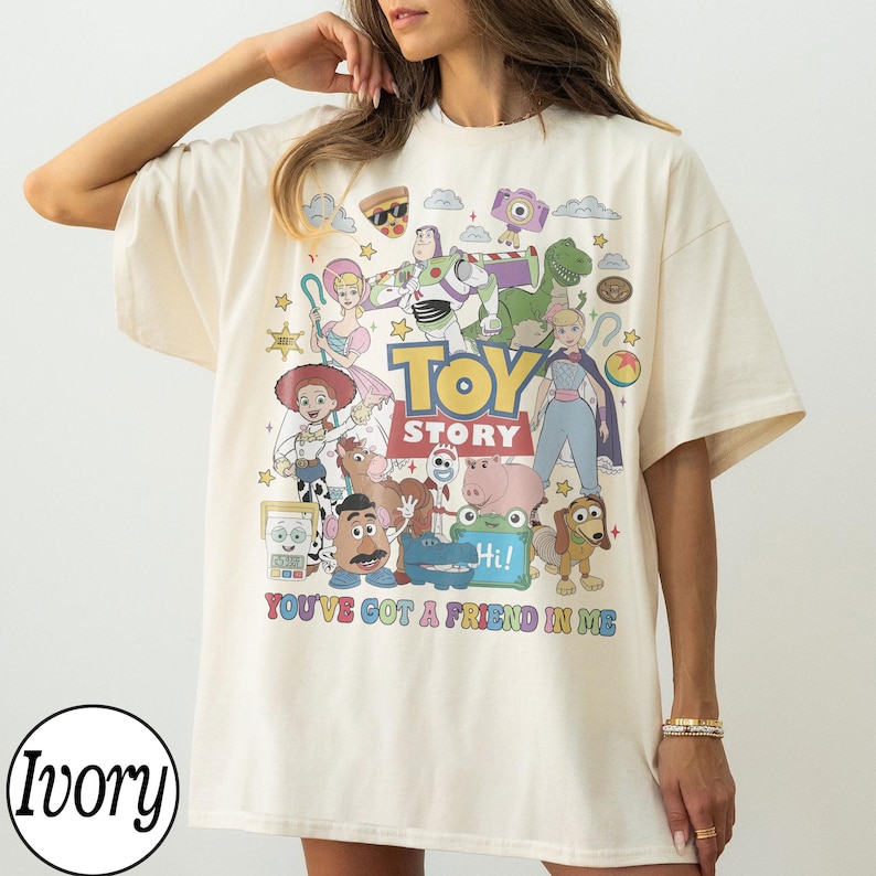 May include: An ivory t-shirt featuring a colorful graphic print of Toy Story characters including Buzz Lightyear, Woody, Jessie, and others. The text "TOY STORY" is centered above the phrase "YOU'VE GOT A FRIEND IN ME" in multi-colored letters.