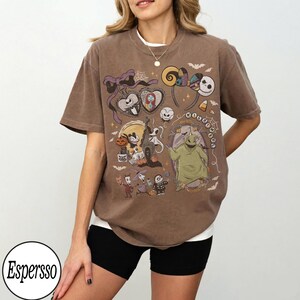 May include: Brown t-shirt featuring a colorful graphic of characters and elements from "The Nightmare Before Christmas." The design includes Jack Skellington, Sally, Oogie Boogie, and other iconic imagery. The shirt has a relaxed fit and short sleeves.