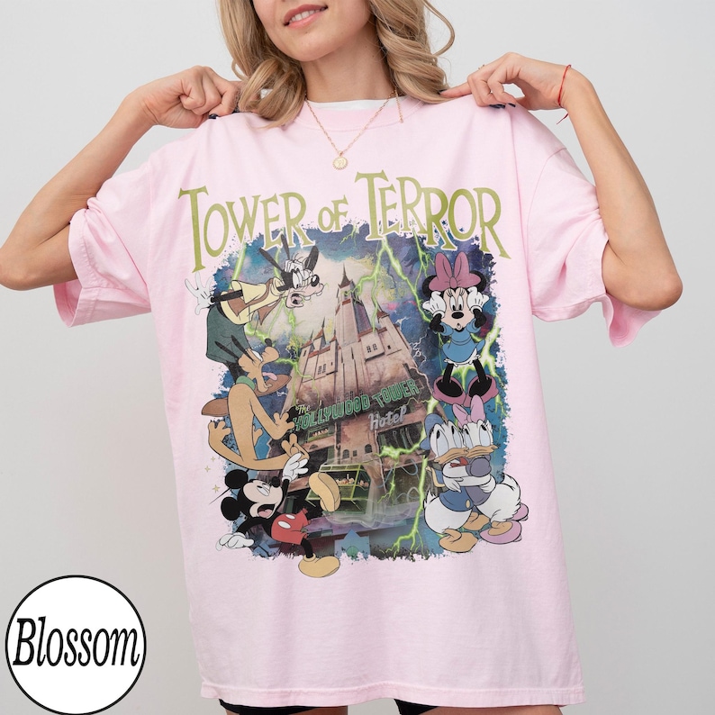 K&ouml;nnte beinhalten: Hellrosa T-Shirt mit dem Schriftzug "Tower of Terror" in Gr&uuml;n und einer Grafik des Hollywood Tower Hotels und Disney-Figuren. Das Design zeigt Micky Maus, Minnie Maus, Goofy, Donald und Daisy Duck.